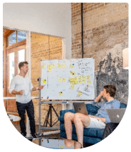 One man in front of a whiteboard explainig graph to a man sitting in front of a him